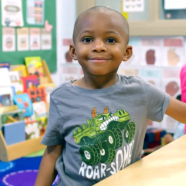 A young child wearing a gray "Roar-some" t-shirt smiles in a classroom setting with books and colorful decorations in the background.