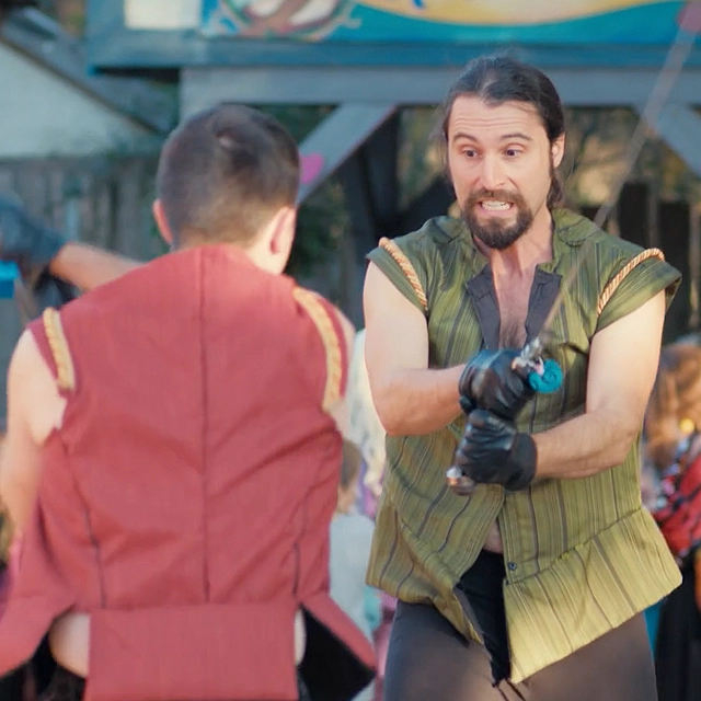 Two men in period costumes are fencing with swords outdoors. The man on the right appears focused, while the man on the left has his back turned to the camera.