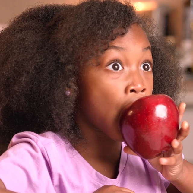 A young girl with curly hair and a surprised expression holds a large red apple up to her mouth, preparing to take a big bite.