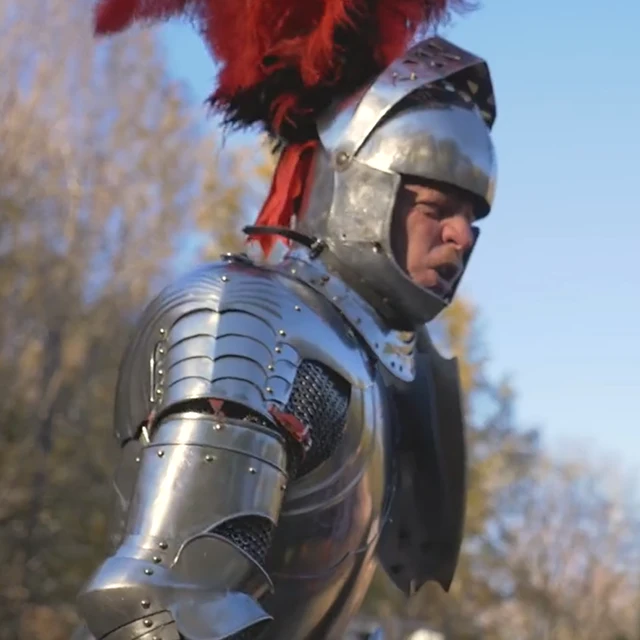 A person in shiny medieval armor and a helmet with red feathers stands outdoors, appearing to shout or speak, with trees in the background.