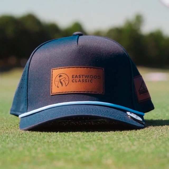 A navy blue baseball cap with a brown leather patch labeled "Eastwood Classic" sits on grass, with another cap blurred in the background.