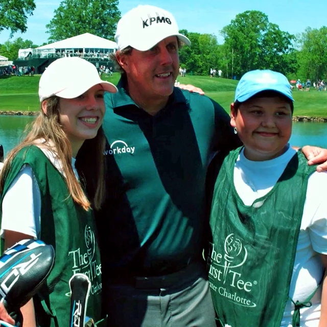 A man in a dark golf shirt and cap stands with two smiling young people in green caddie bibs at a golf course, with trees and a tent in the background.