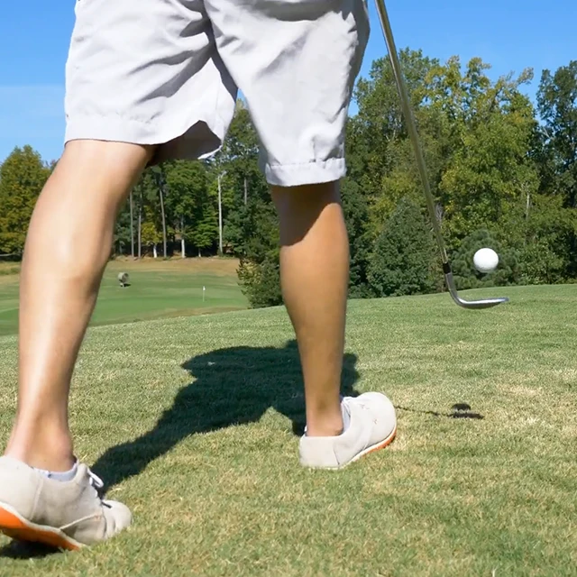 A person in shorts stands on a golf course, swinging a golf club to hit a golf ball into the air. Trees and a blue sky are visible in the background.